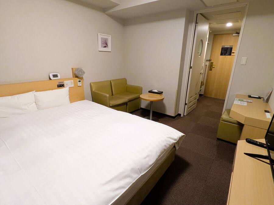 A hotel room with a bed, a desk, a chair, a small table, a TV, and a door leading to another room.