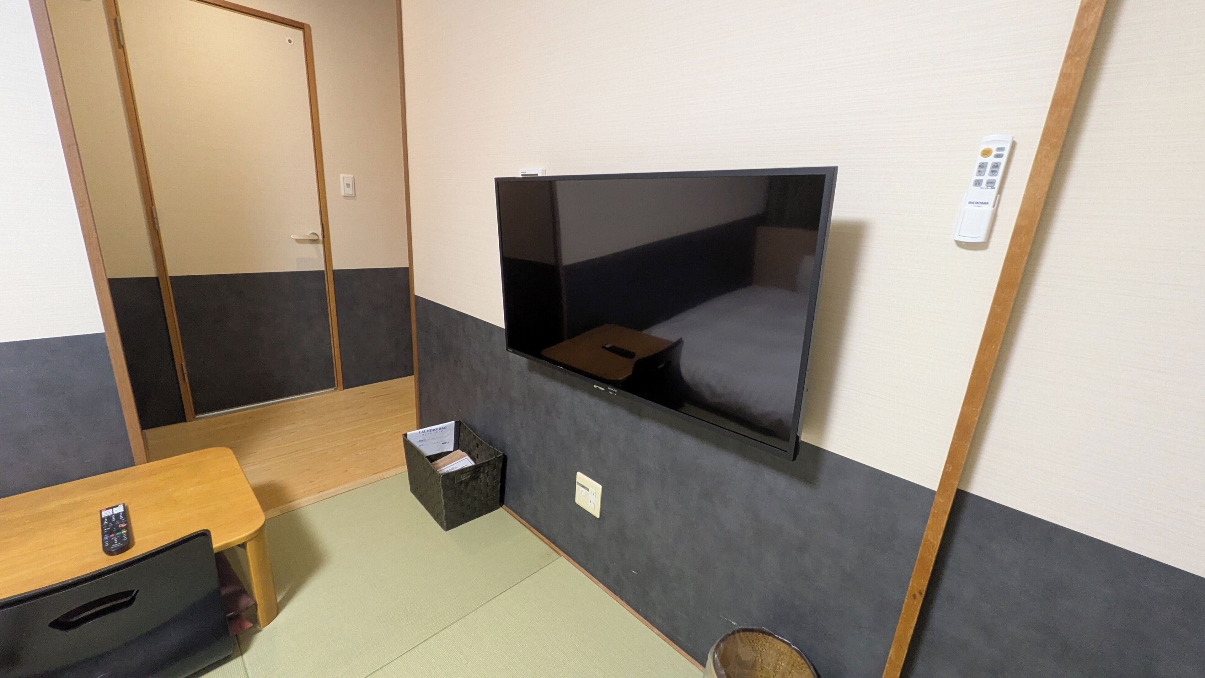 Una habitación con un televisor montado en la pared, un escritorio de madera con un control remoto, una mesita con un control remoto y un cubo de basura negro.