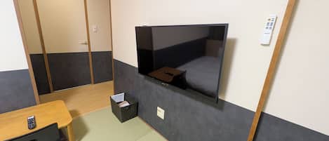 Una habitación con un televisor montado en la pared, un escritorio de madera con un control remoto, una mesita con un control remoto y un cubo de basura negro.
