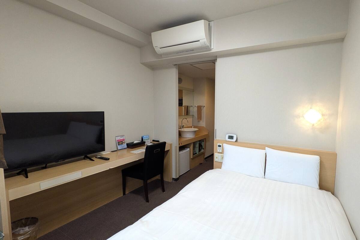 A hotel room with a bed, desk, chair, television, and an air conditioning unit.