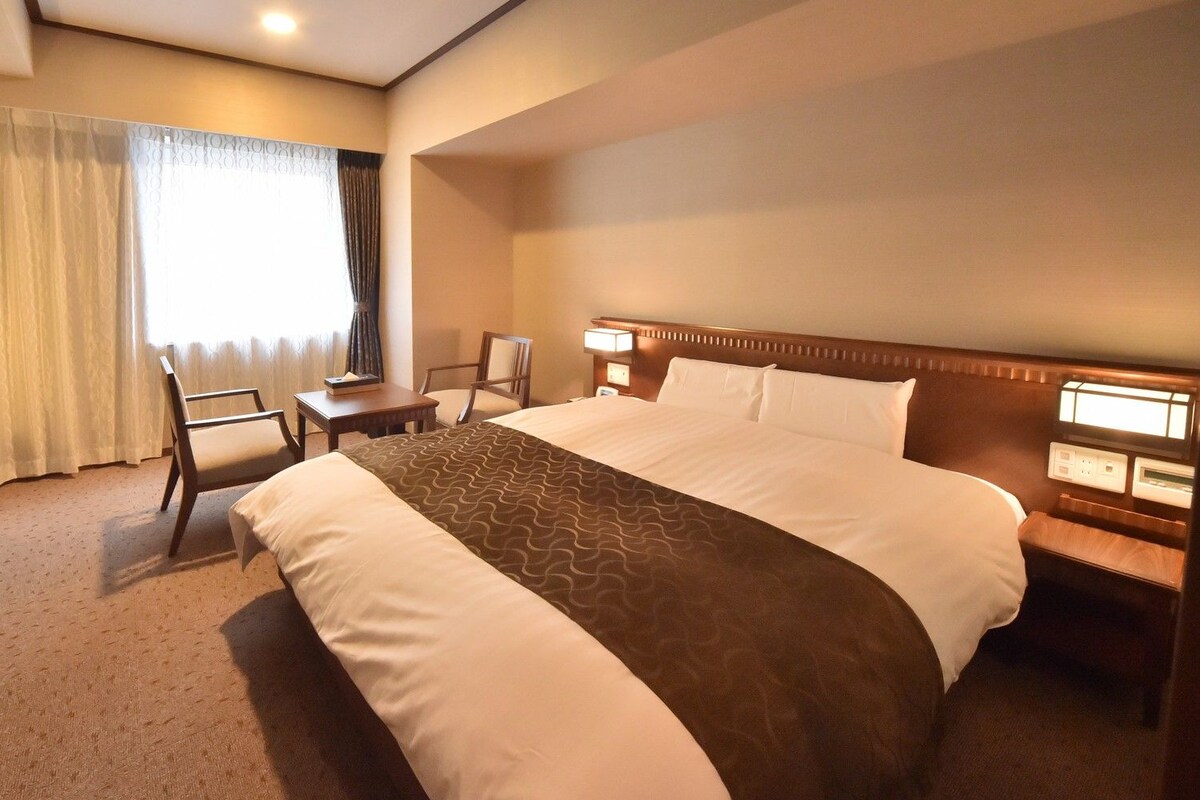 A hotel room with a large bed, a desk, a chair, and a nightstand with a lamp.