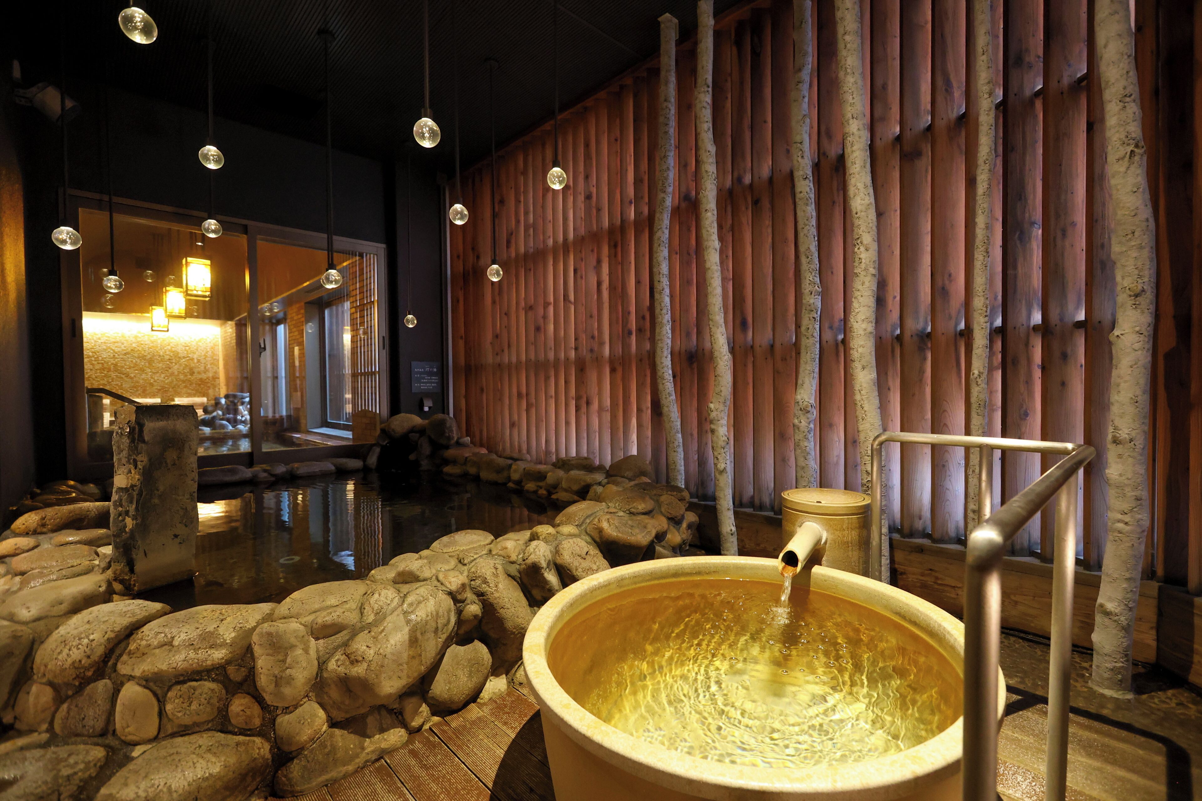 A hot spring bath area with a wooden wall, stone rocks, and a large wooden tub.