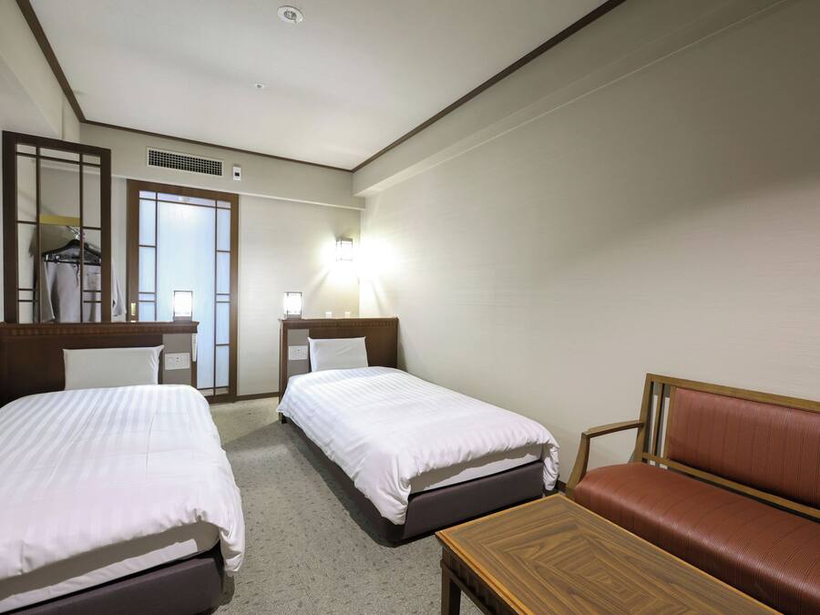 Ein Hotelzimmer mit zwei Einzelbetten, einem Holfnachtkästchen, einem braunen Ledersofa, einem Holztisch, einem Spiegel, einer Wandlampe und einer Tür mit Fenster.
