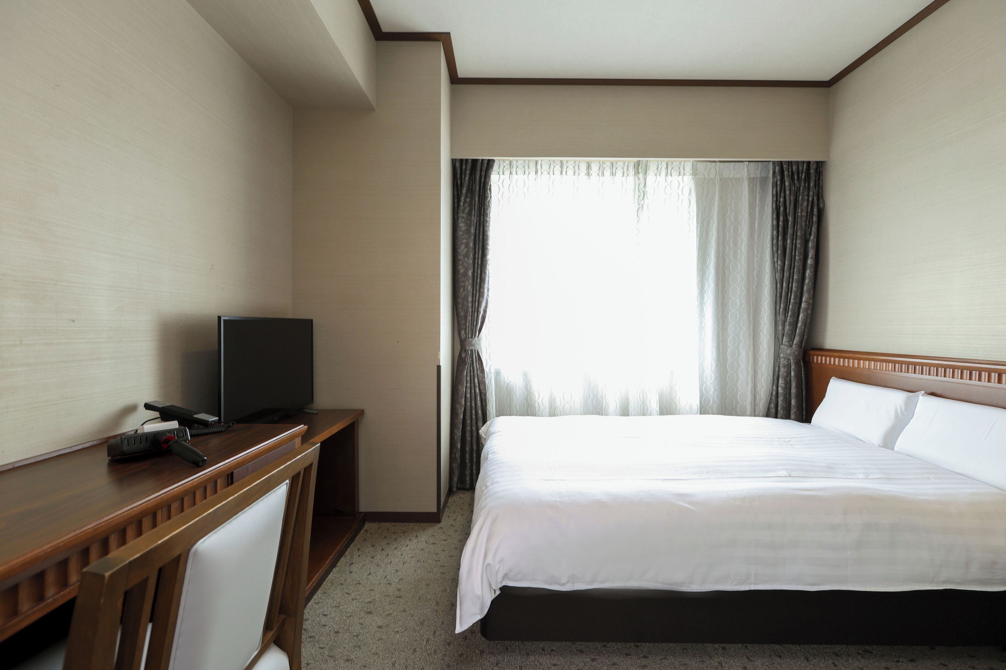 A hotel room with a bed, a desk with a television, and a window with curtains.