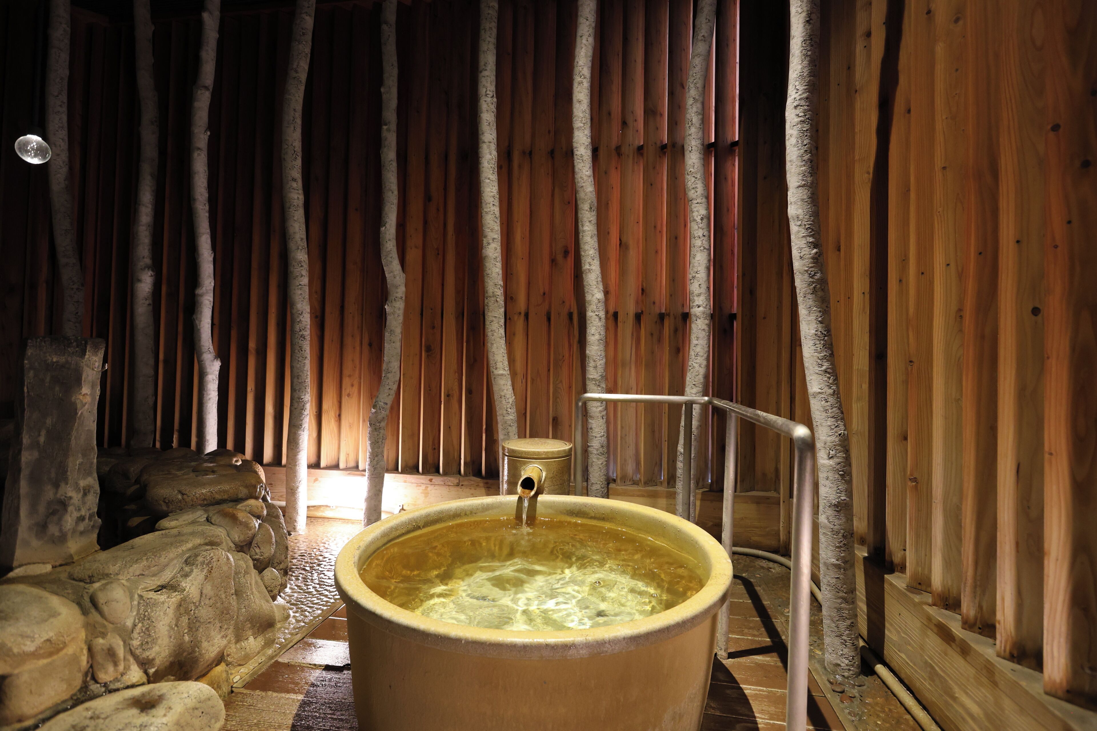 Un intérieur en bois avec un grand bassin en pierre rempli d’eau, une rambarde en métal et un banc en bois.