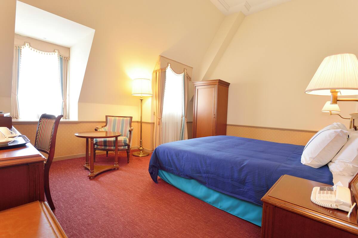 A hotel room with a bed, a desk, a chair, a lamp, and a telephone.