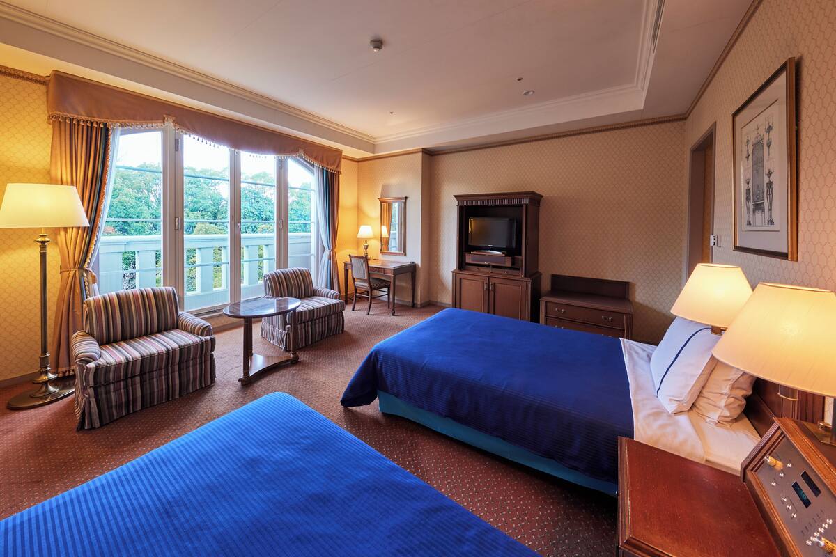 A hotel room with two beds, a TV, a desk, and a balcony with curtains.