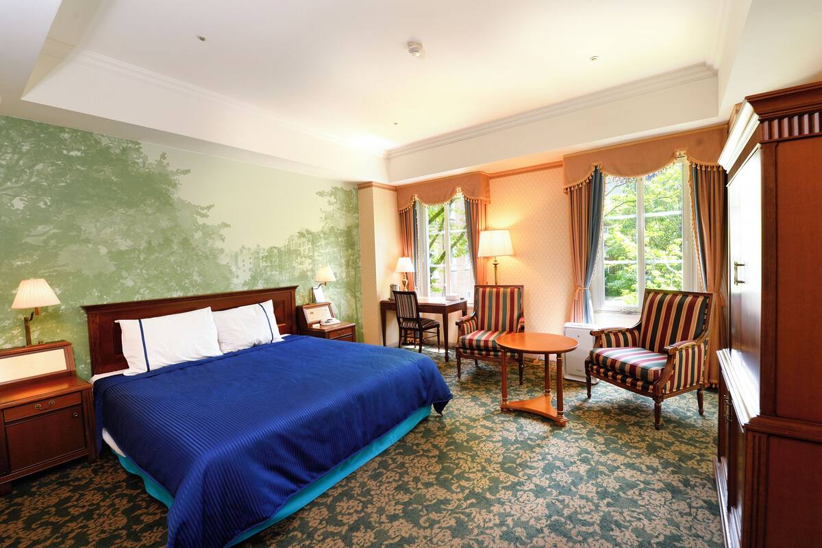 A hotel room with a large bed, a desk, two chairs, a lamp, and a view of greenery through the window.
