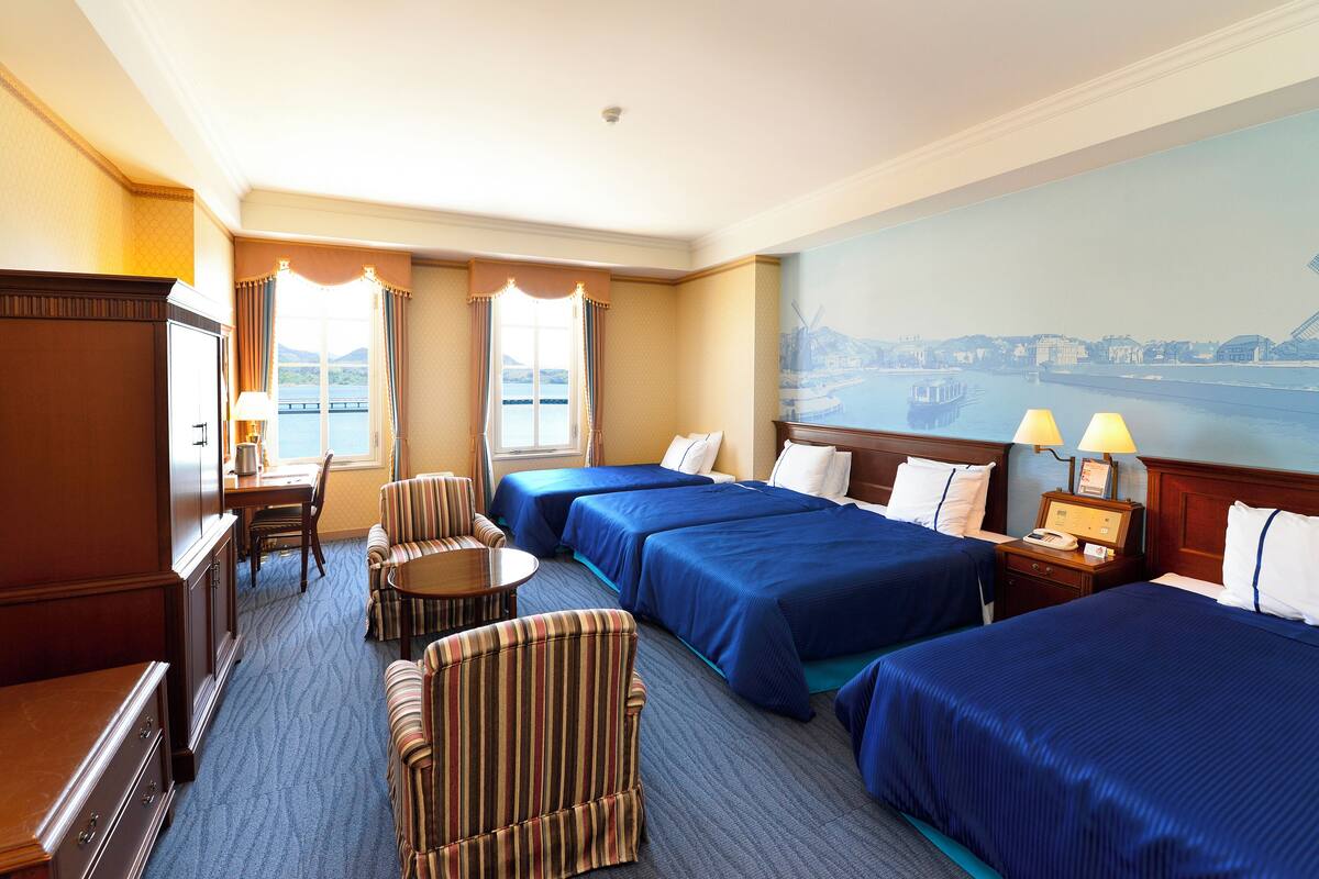 A hotel room with three beds, a dining table, a striped armchair, a desk, and a large window with a view of a body of water and buildings.