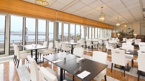 A spacious dining area with tables and chairs, large windows offering a view of the sea, and a wooden ceiling. - Hotel Den Haag Huis Ten Bosch (Sasebo)