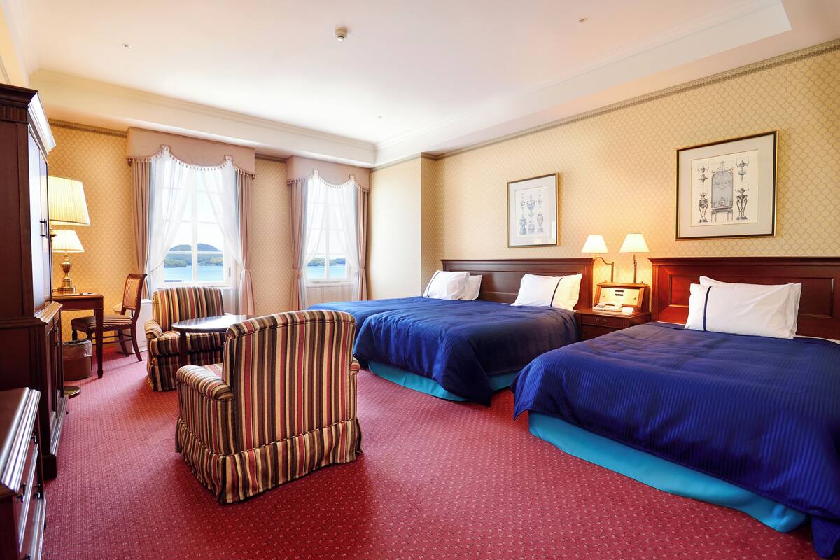 A hotel room with two beds, a striped armchair, a small table, and a view of the sea.