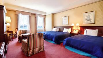 A hotel room with two beds, a striped armchair, a small table, and a view of the sea.