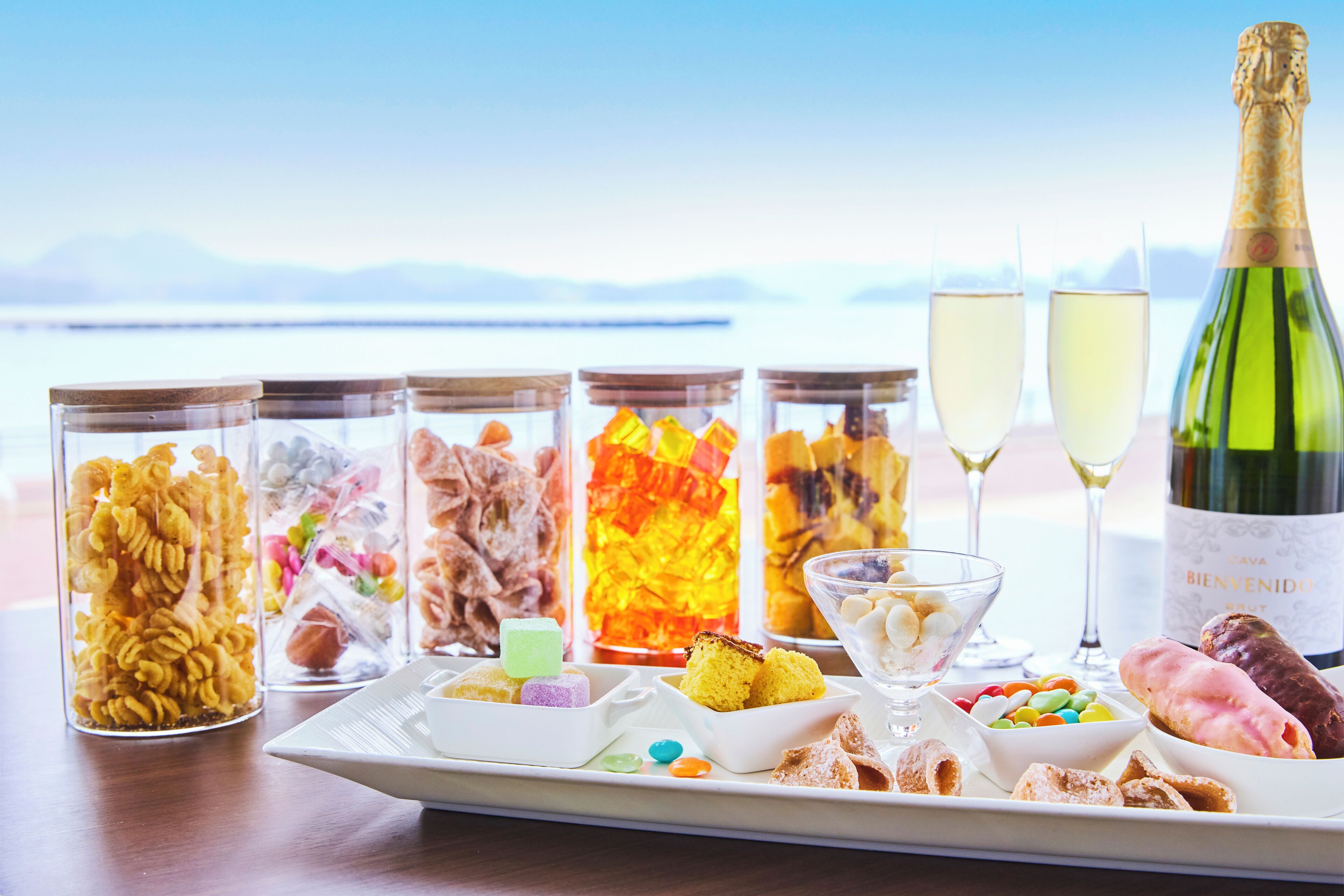 A table with various food items and two glasses of champagne, set against a scenic backdrop.