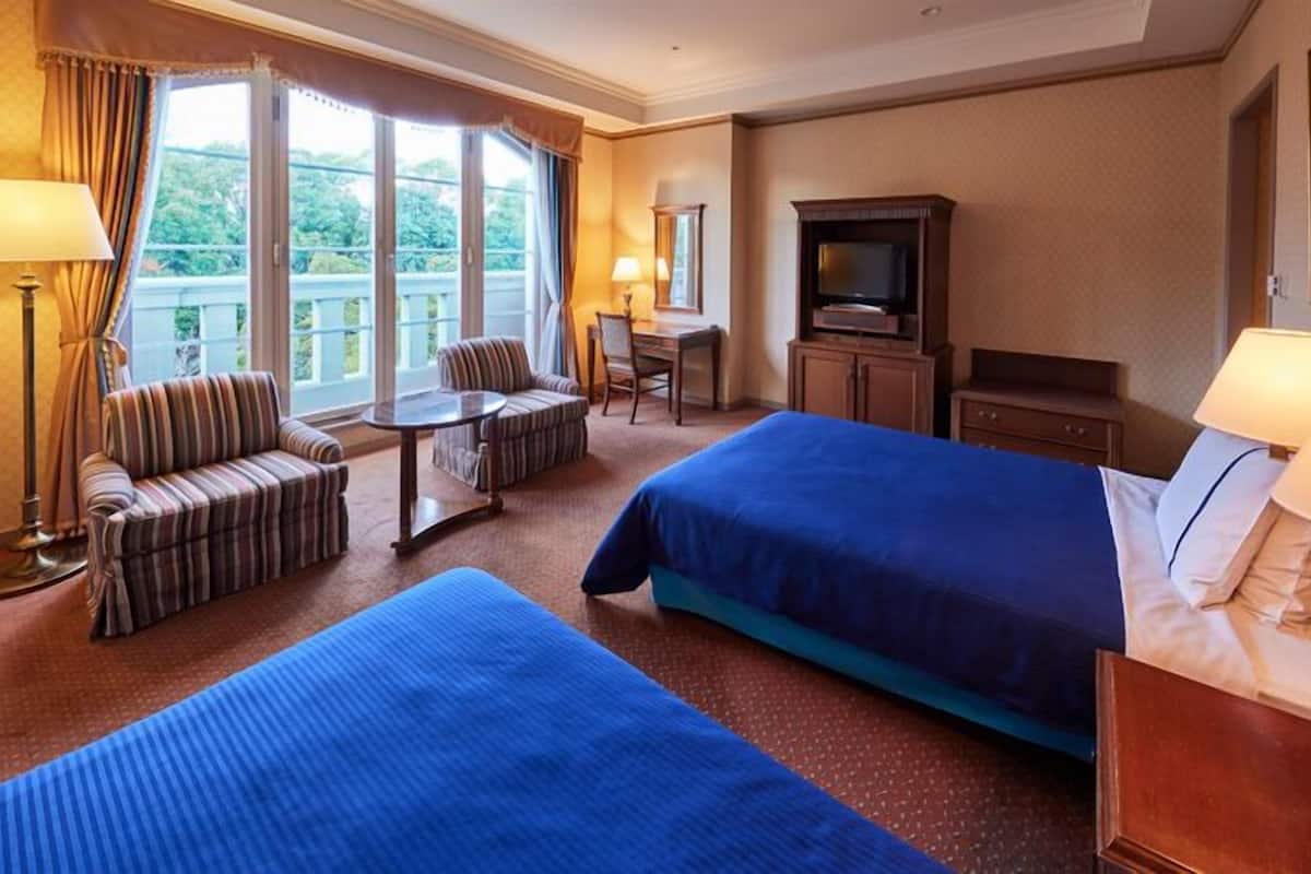 A hotel room with two beds, a sofa, a small table, a TV, and a balcony with curtains.