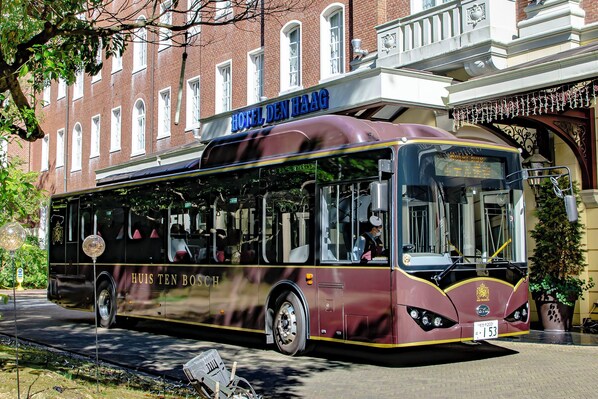 Un autobus de couleur bordeaux, sur lequel on pouvait lire les inscriptions « HUIS TEN BOSCH » et « HOTEL DEN HAAG », était garé devant un immeuble orné d’une enseigne.