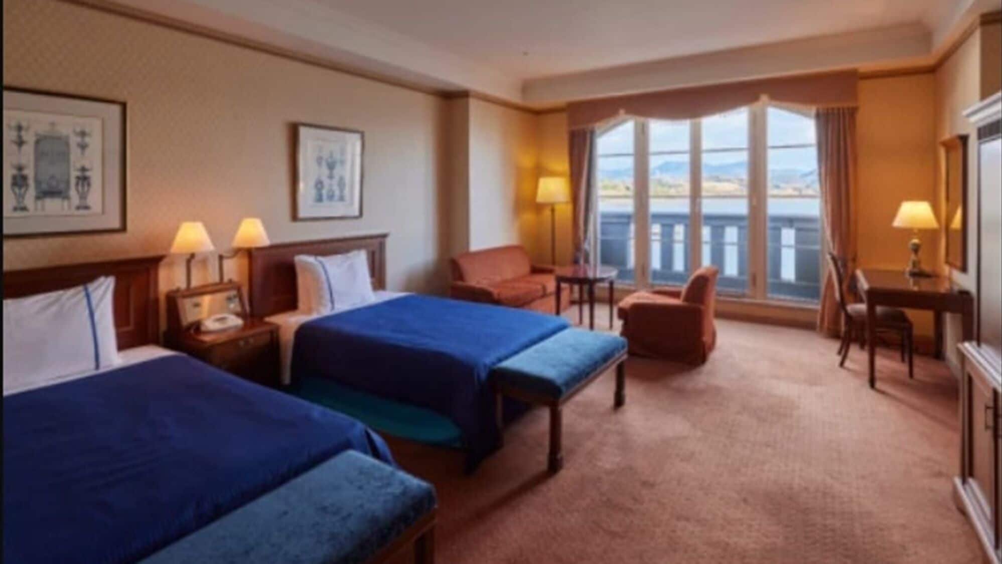 A hotel room with two beds, a desk, a chair, a lamp, and a large window with a view.