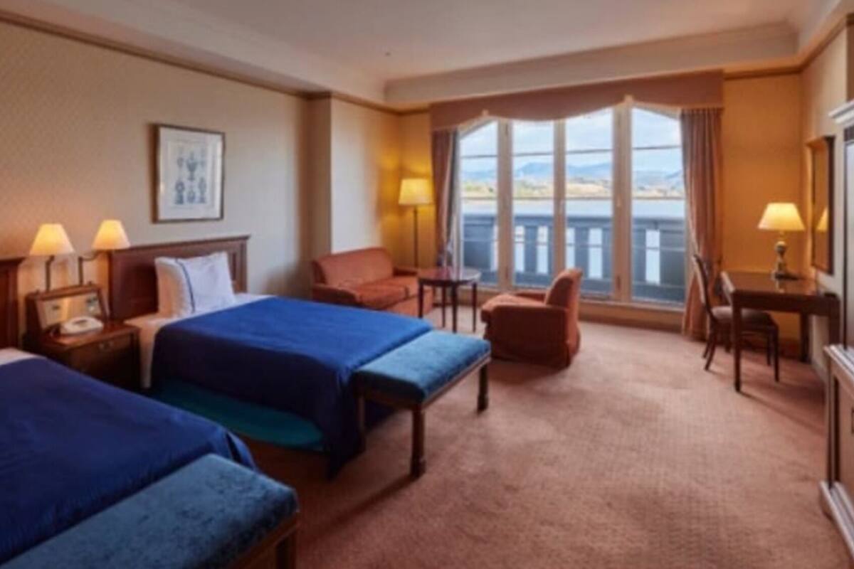 A hotel room with two beds, a desk, a chair, a lamp, and a large window with a view.