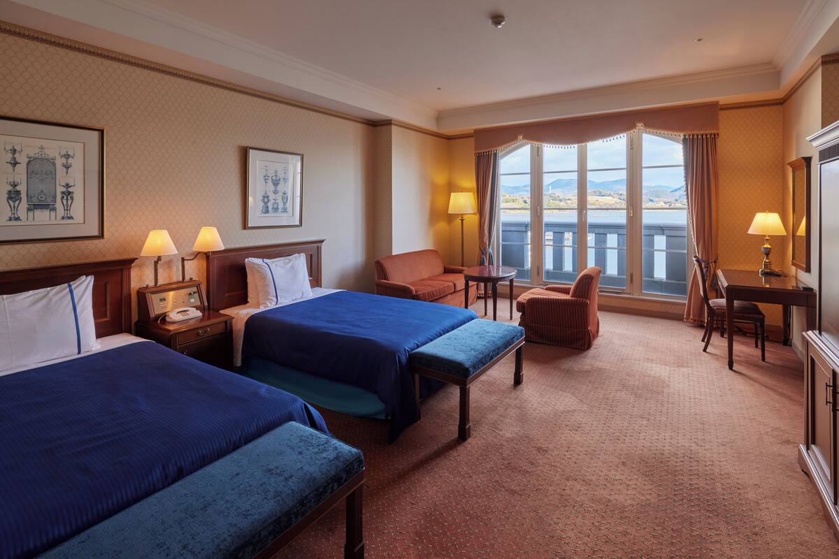 A hotel room with two beds, a desk, a chair, and a view of the outdoors.