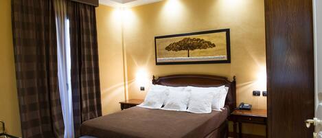Habitación de hotel con cama, escritorio, silla, mesita de noche, lámpara y un cuadro en la pared.