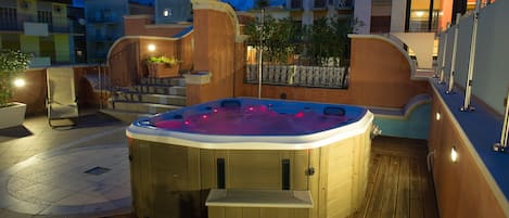 A hot tub in a courtyard with steps leading up to it, surrounded by buildings and potted plants.