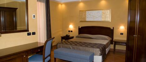 A hotel room with a bed, bedside tables, a desk with a chair, a bench, a large mirror, and a wall-mounted artwork.
