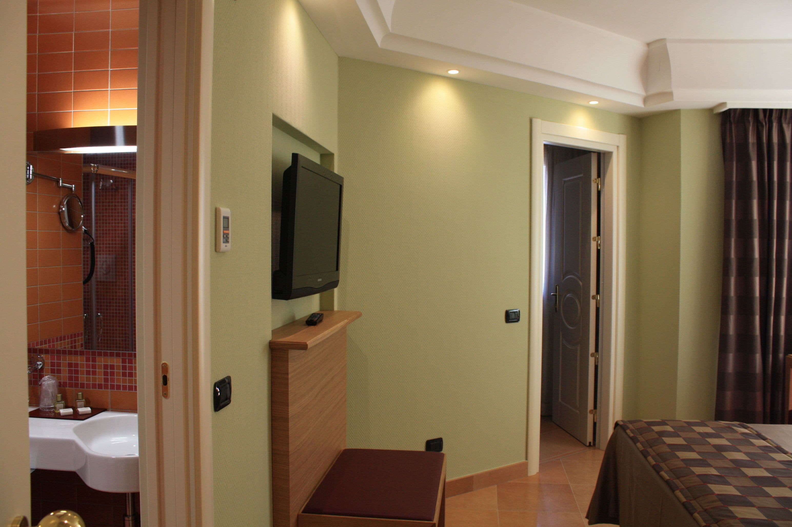Habitación de hotel con televisión, un pequeño banco de madera, una puerta y un baño visible a través de una puerta abierta.