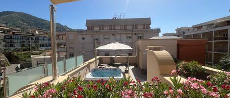 A rooftop terrace with a hot tub, outdoor seating, and a view of a residential area with a mountain in the background.