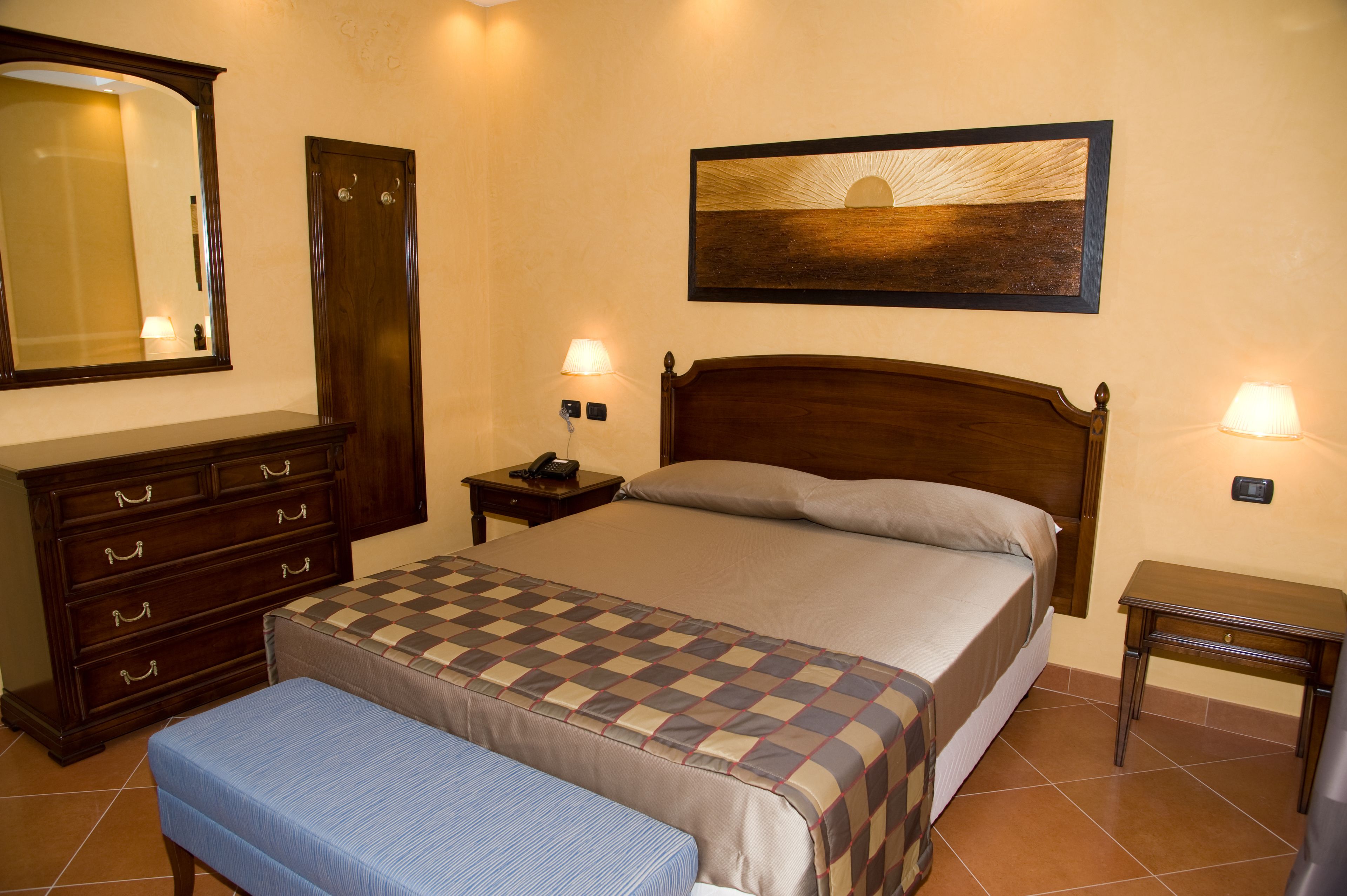 A bedroom with a bed, a dresser, a bench, a nightstand, a wall-mounted lamp, and a painting.