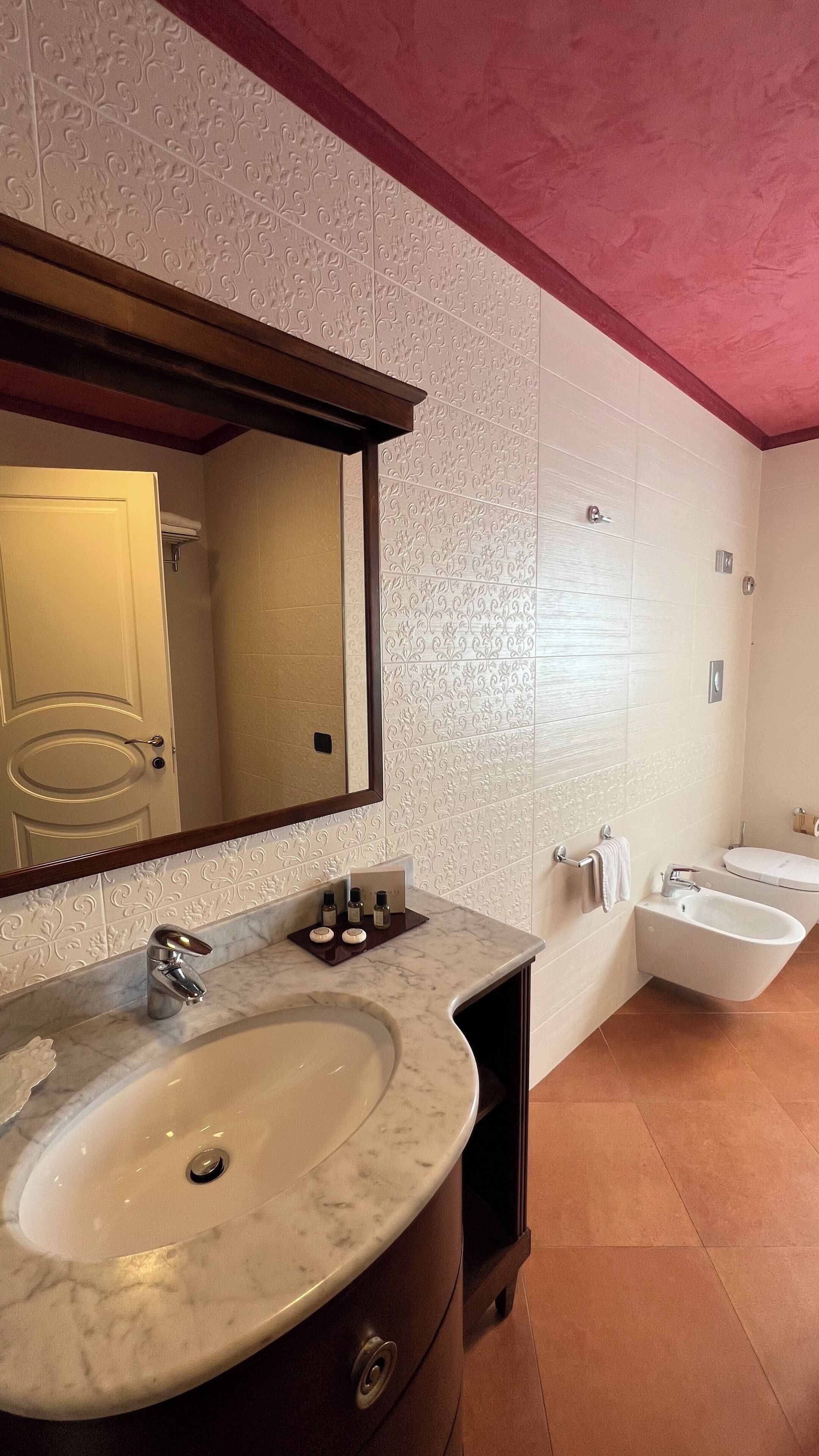 Un baño con lavamanos de mármol, un espejo grande, dos inodoros y un perchero para toallas.