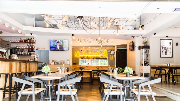 A modern restaurant interior with wooden tables and chairs, a bar area with bottles, and a television screen. - Vela Boutique Hotel (Hong Kong)