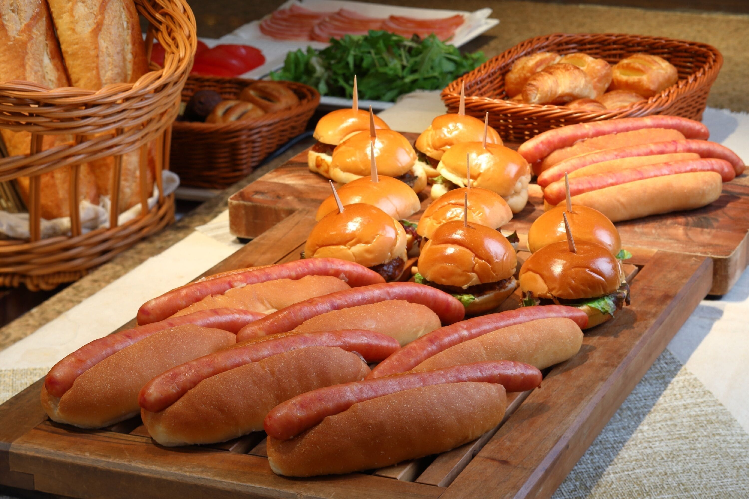 A wooden table with hot dogs and buns, baskets of bread, and a plate of vegetables.