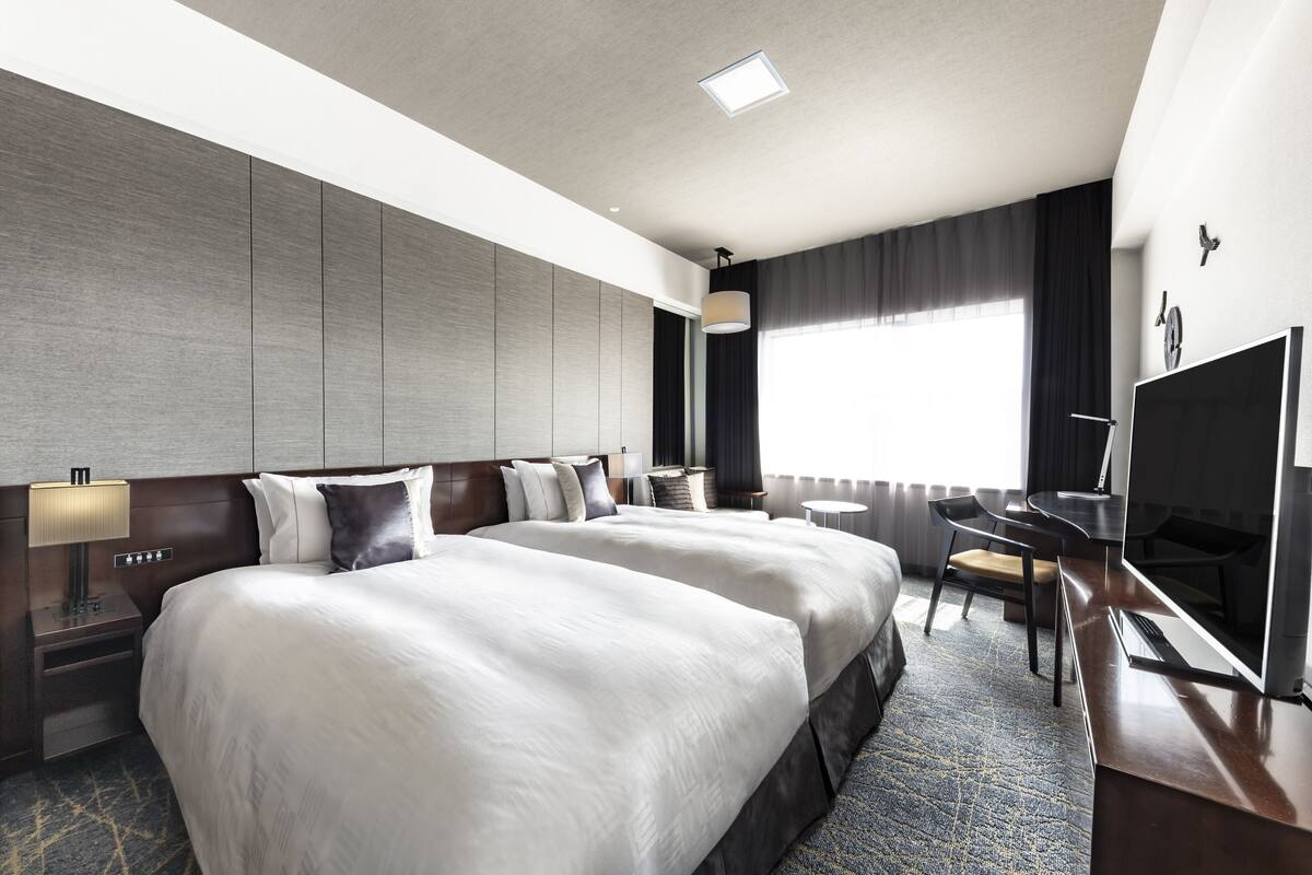 A hotel room with two large beds, a desk with a TV, and a window with curtains.