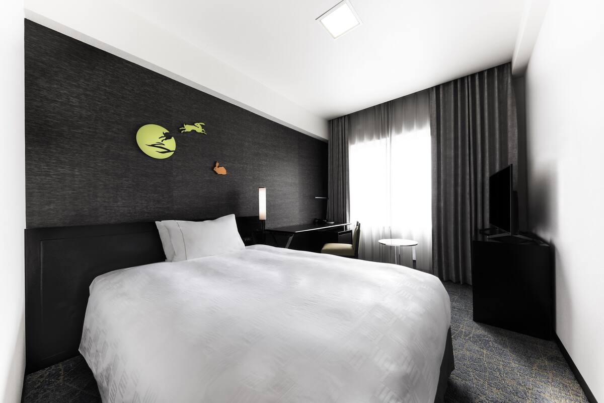 A modern hotel room with a large bed, a desk, a chair, a TV, and a wall with a logo.