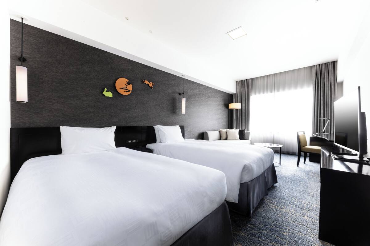 A hotel room with two beds, a desk, a chair, and a TV.