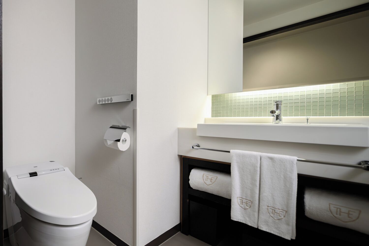 A modern bathroom with a toilet, a paper towel dispenser, a towel rack, and a sink with a mirror above it.