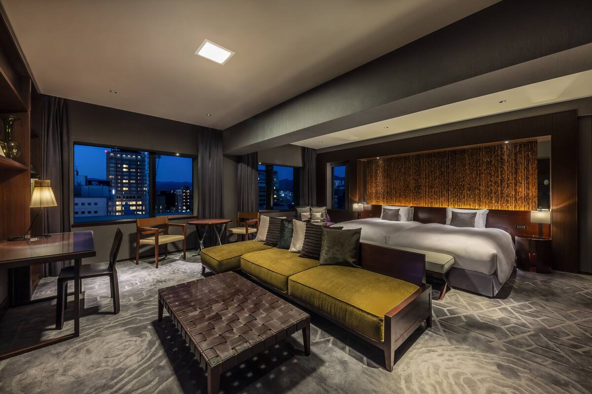 A modern hotel room with a large bed, a sofa, a desk, and a view of the city at night.