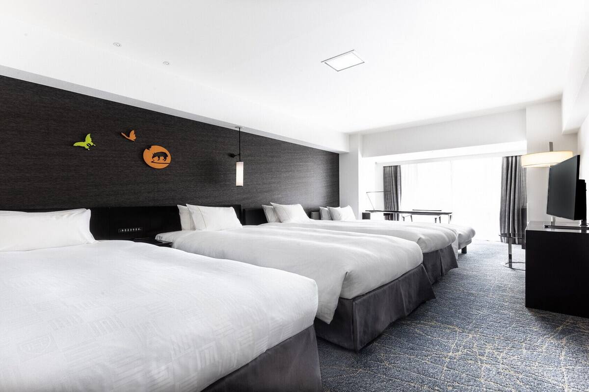 A hotel room with three beds, a desk, a TV, and a large window.