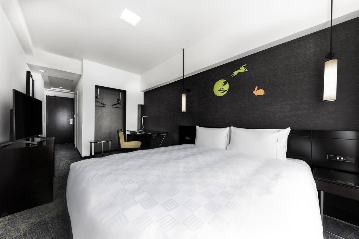 A modern hotel room with a large bed, a desk, and a chair. There is a TV on the left side and a wardrobe. The room has a dark wall with a circular design and a hanging light fixture.