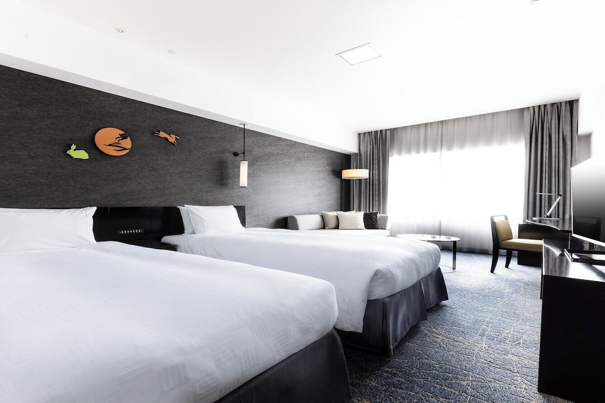 A hotel room with two beds, a desk, a chair, a TV, and a large window with curtains.