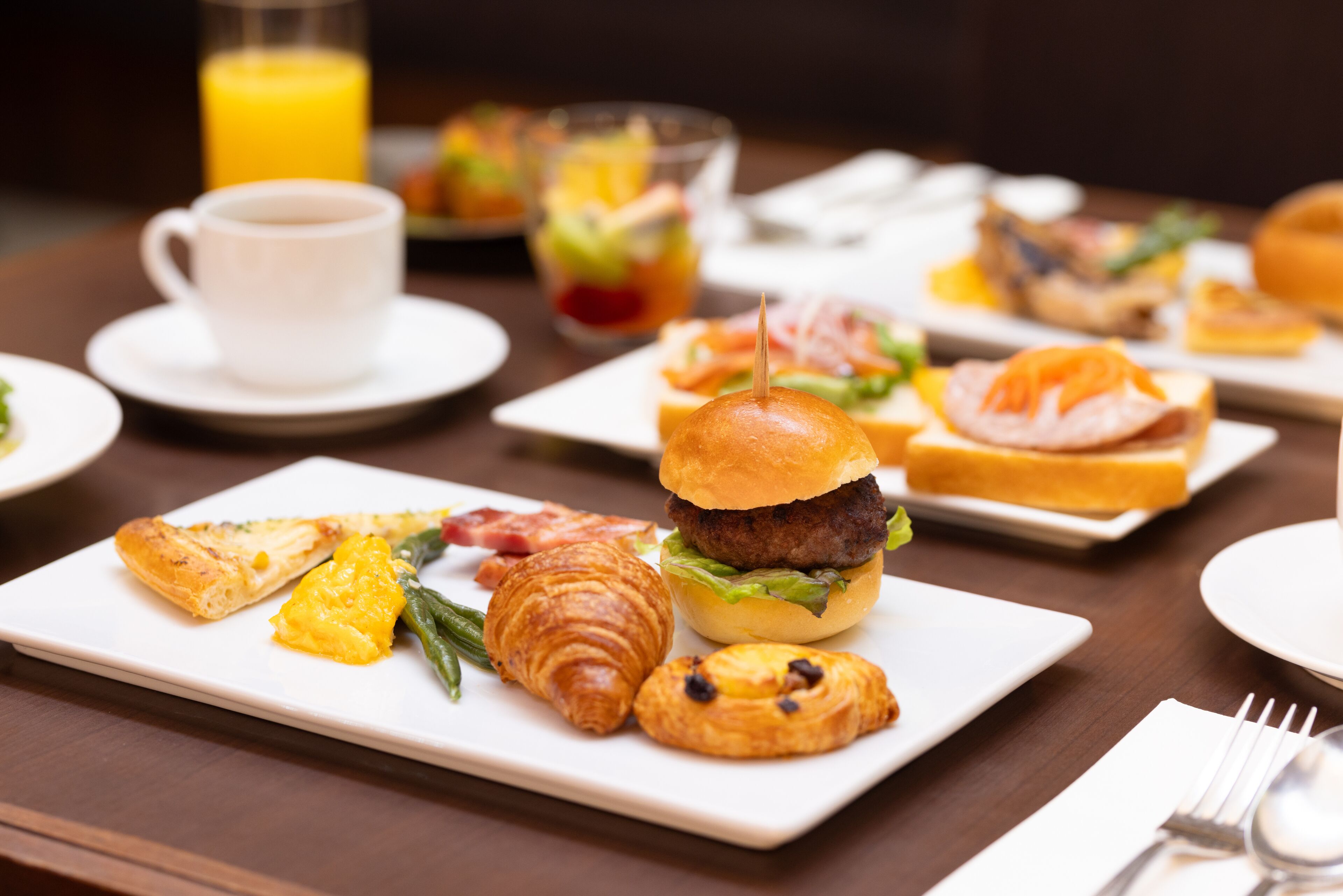 Un bufé de desayuno con diversos platos, incluyendo croissants, una hamburguesa y productos de pastelería.