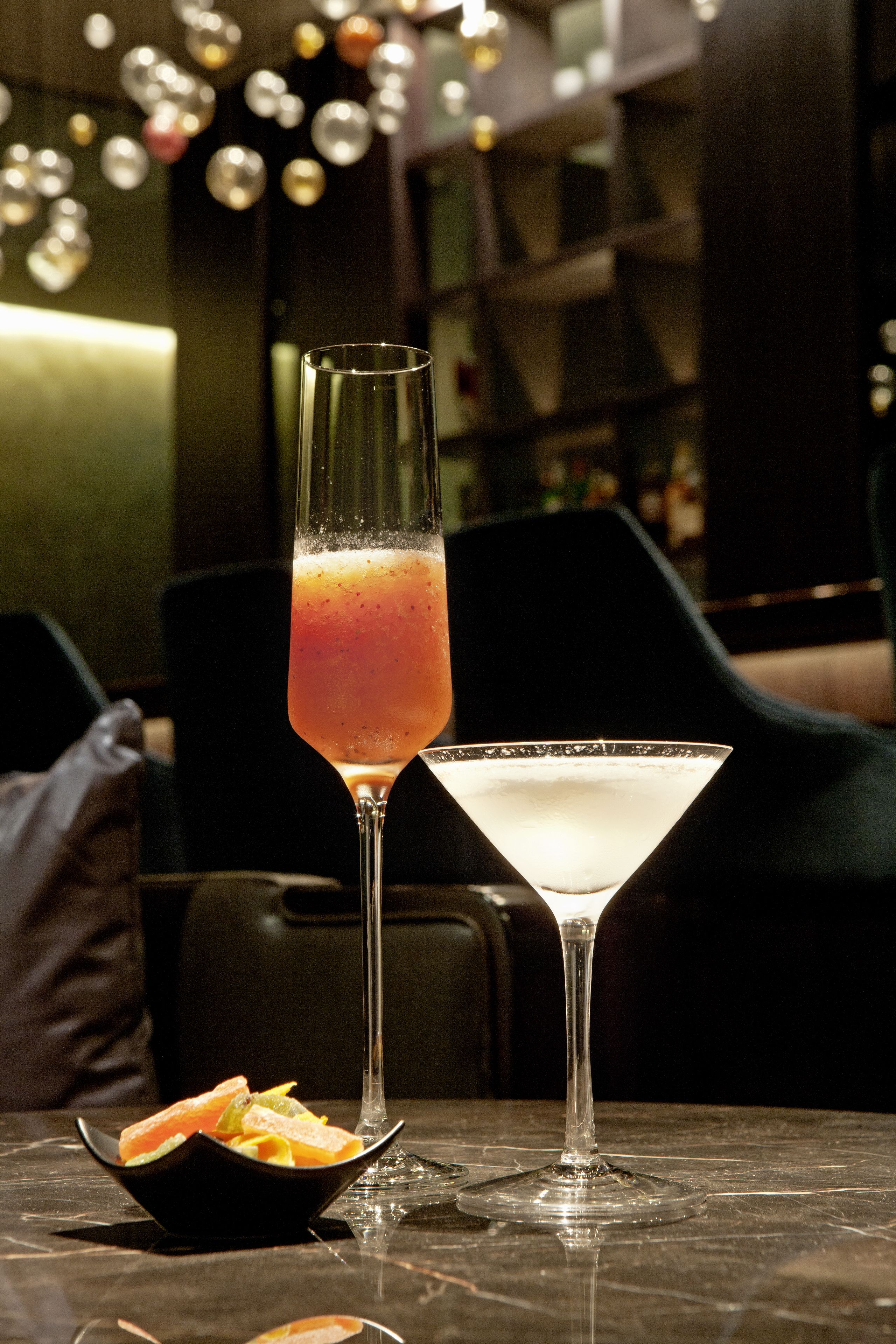 Dos vasos de cóctel y un bol de fruta sobre una mesa en un bar con poca luz.