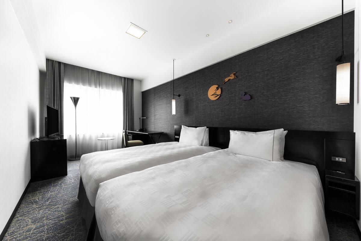 A modern hotel room with two large beds, a desk, a chair, and a large window with curtains.