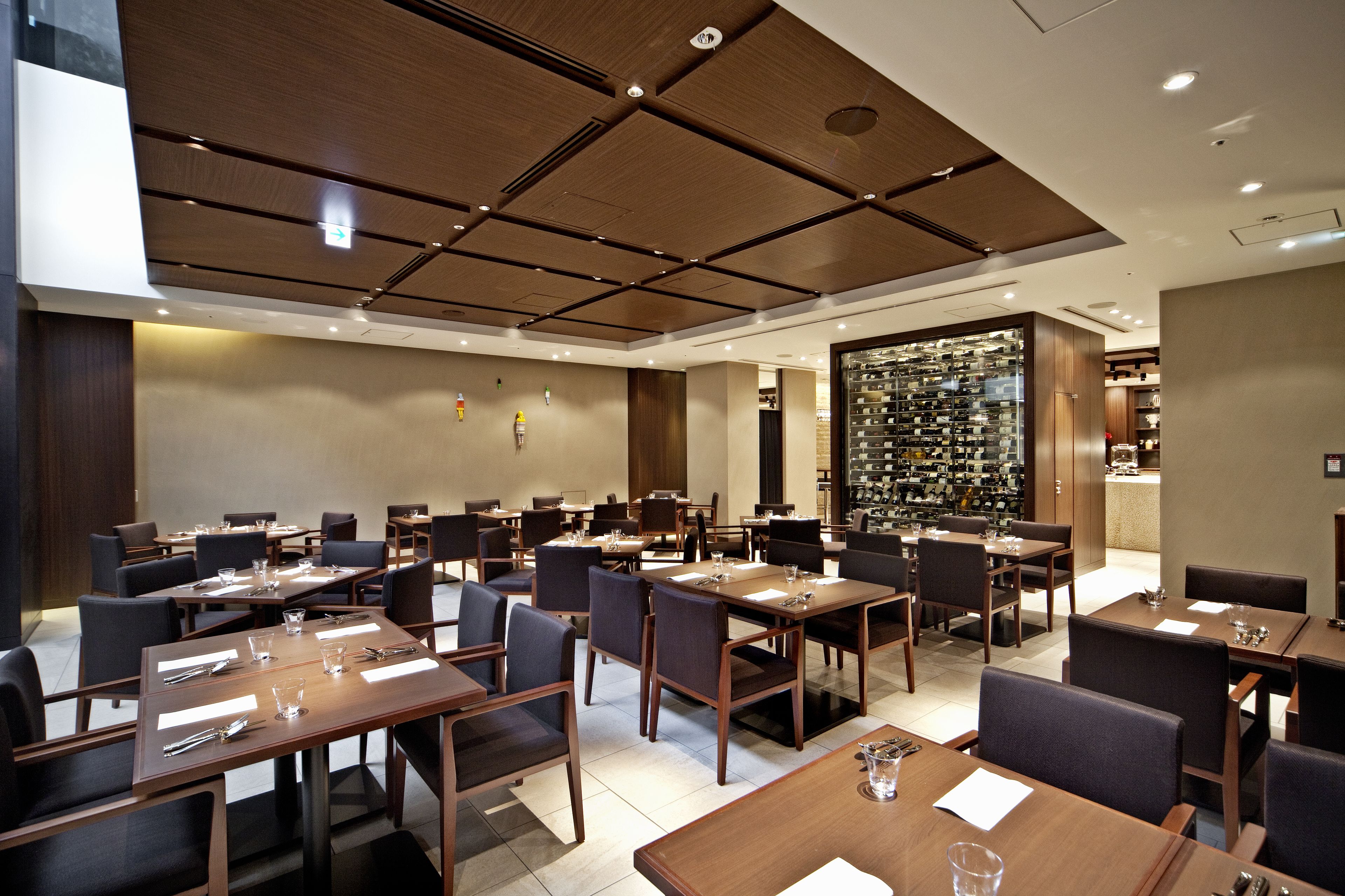 A modern hotel dining area with multiple tables and chairs, a wine rack, and a view of the kitchen.