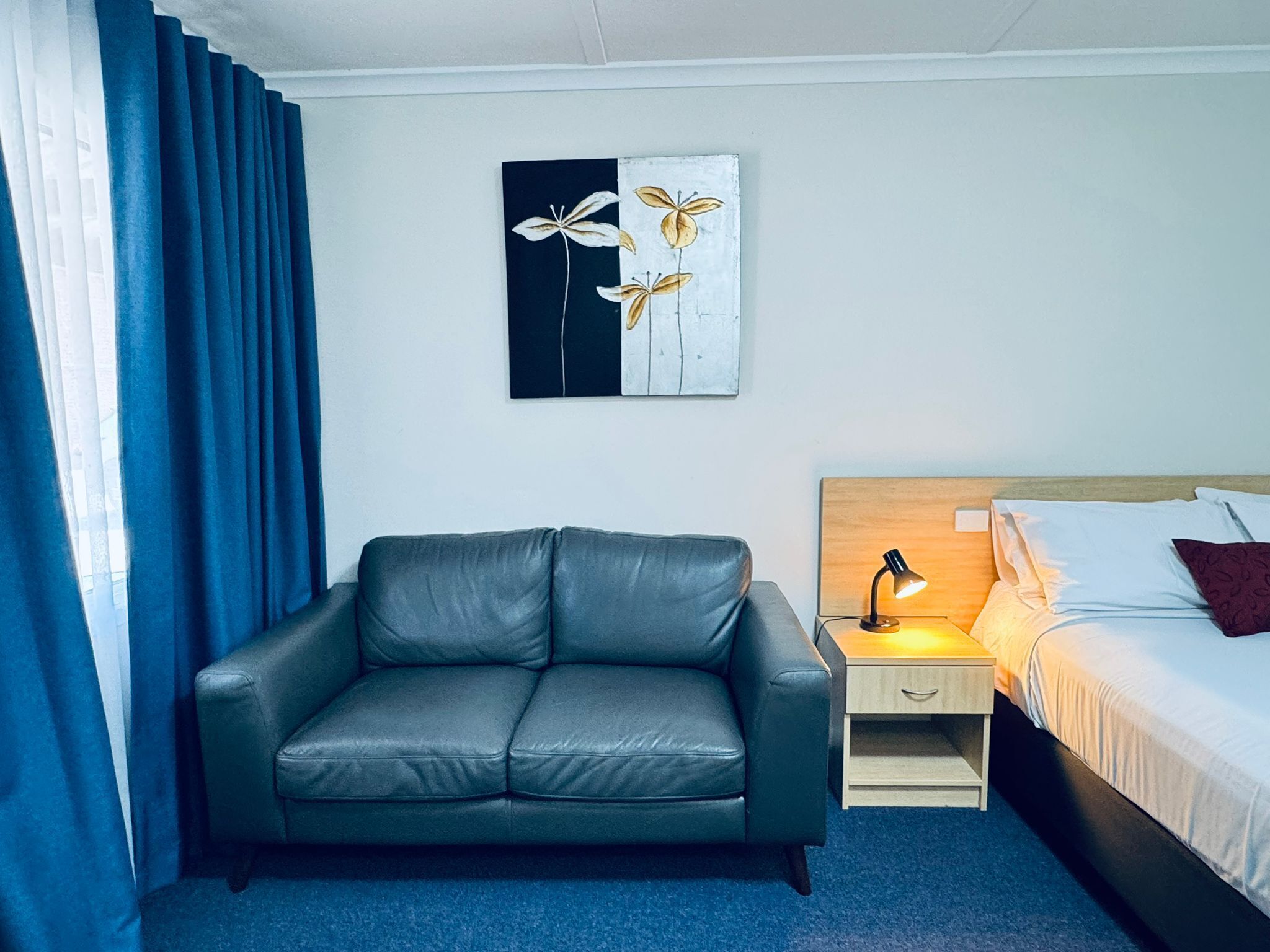 A room with a grey leather sofa, a bed with white bedding and a red pillow, a nightstand with a lamp, and a wall-mounted artwork.