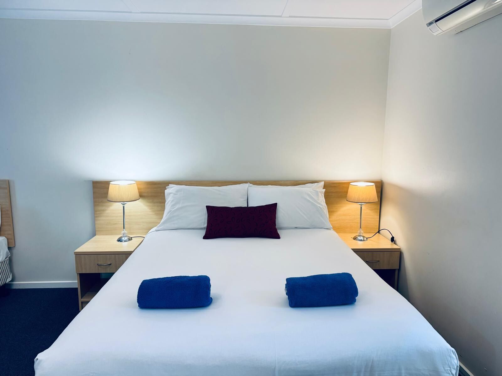 A hotel room with a bed, two nightstands, a wooden headboard, a single red pillow, two blue towels, and two lamps.