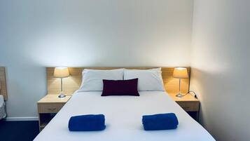 A hotel room with a bed, two nightstands, a wooden headboard, a single red pillow, two blue towels, and two lamps.