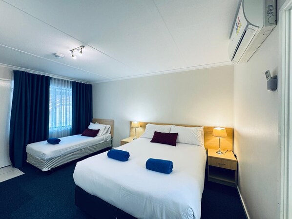 A hotel room with two beds, a window with curtains, a bedside table with a lamp, and an air conditioning unit.