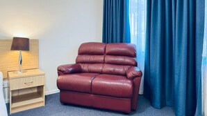 A room with a leather sofa, a wooden nightstand with a lamp, and blue curtains. - Edinburgh Motor Inn (Warragul)