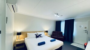 A hotel room with a bed, two bedside tables with lamps, a leather chair, a desk, and a wardrobe. - Edinburgh Motor Inn (Warragul)