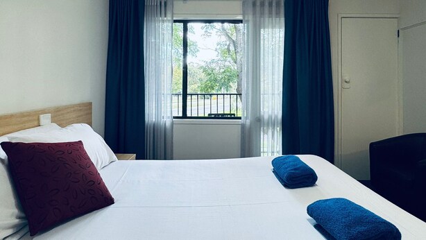 A hotel room with a bed, a window with curtains, a view of the outside, and a door. - Edinburgh Motor Inn (Warragul)